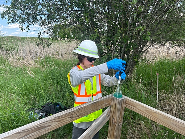 An intern wearing a white hard hat, neon yellow safety vest, and blue latex gloves is seen testing water in a glass container while in a bog