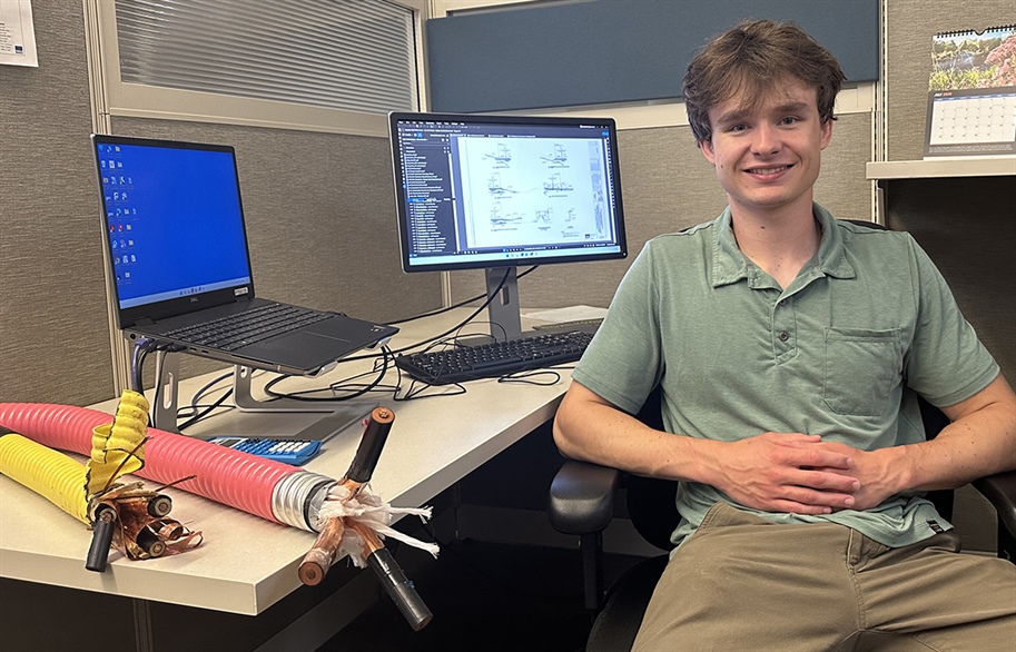 A young man sitting and smiling at an office desk that has two screens and thick electrical wire samples on it.