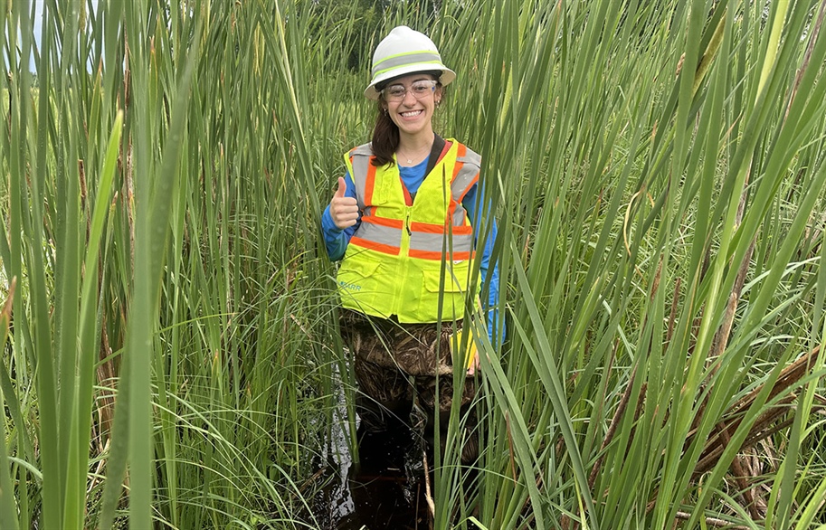 A young women wearing a safety vest and white hard hat is giving a thumbs-up while standing knee deep in a bog surrounded by tall green grasses