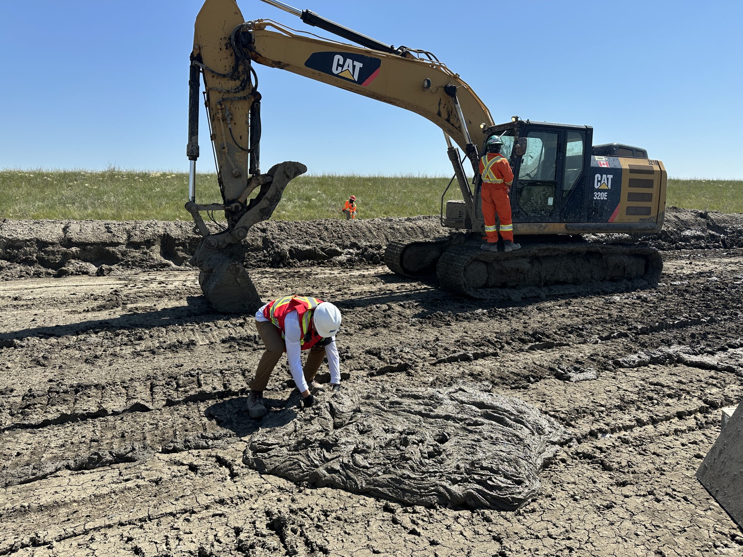 An intern wearing a white hard hat and orange safety vest hovers over a pile of thick slurry backfill to test it with a large machinery vehicle in the background.