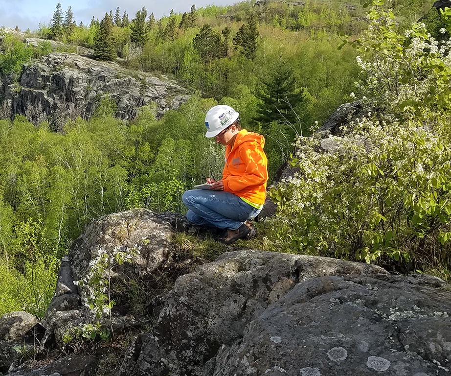a Barr employee wearing a hard hat and bright orange shirt taking field notes on a rocky outcrop