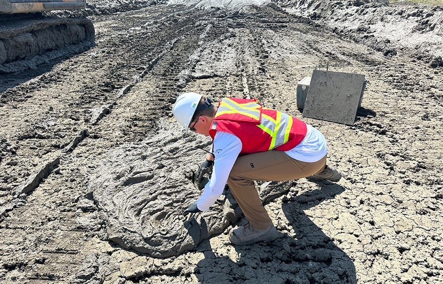 An intern wearing a safety vest and white hard hat hovers over thick slurry backfill on a mine site.