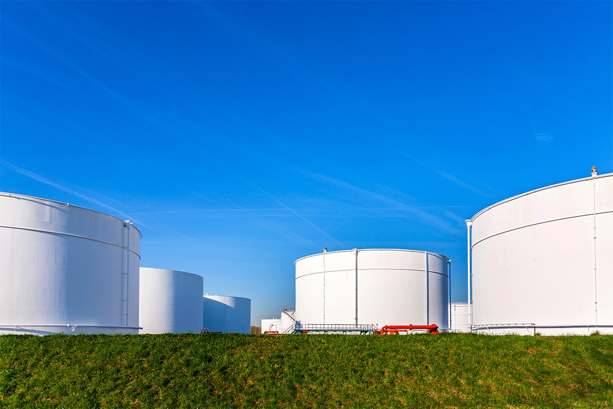 An array of large white fuel storage tanks in a green field under a bright blue sky