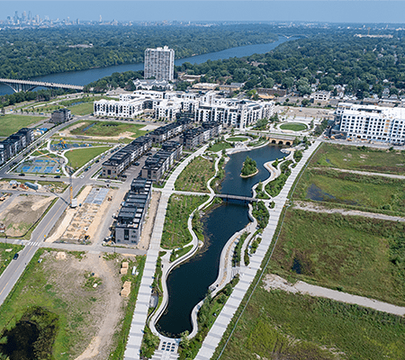 Aerial image of an urban, residential development site next to a large river with a long, narrow pond extending through the middle on a sunny day.