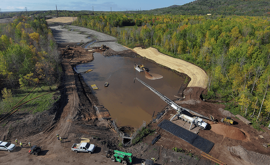 Aerial image of a waterway lined by disturbed soil and construction equipment and surrounded by dense green forest.