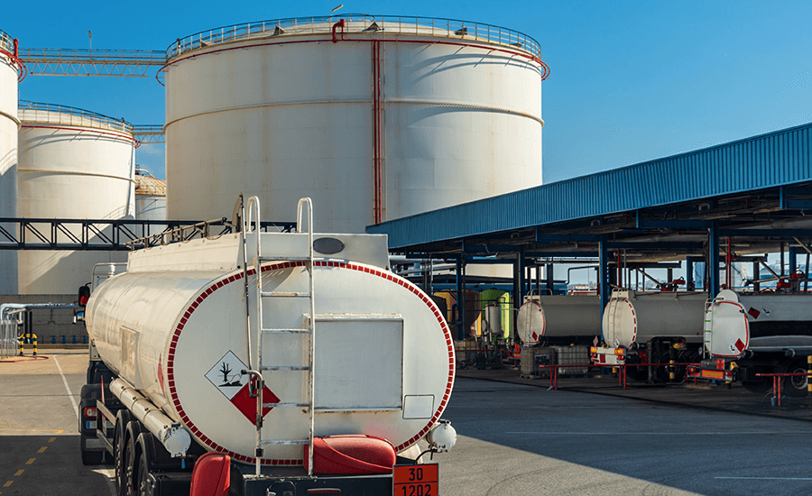 A gasoline tanker truck at a gasoline distribution facility with large storage tanks in the background.