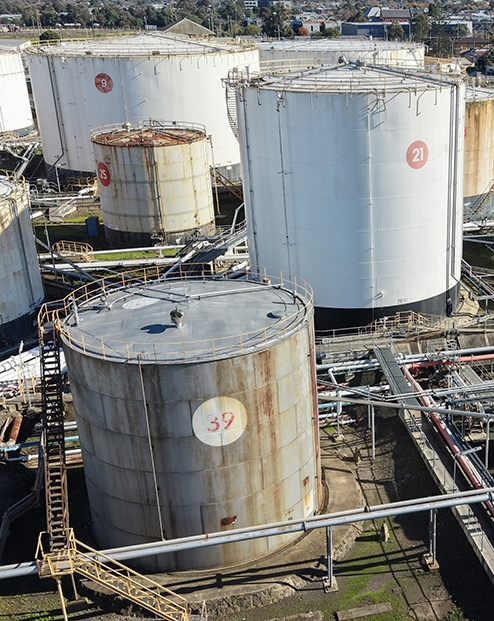 Industrial fuel storage facility with multiple large cylindrical tanks, interconnected pipelines, and processing equipment viewed from above.